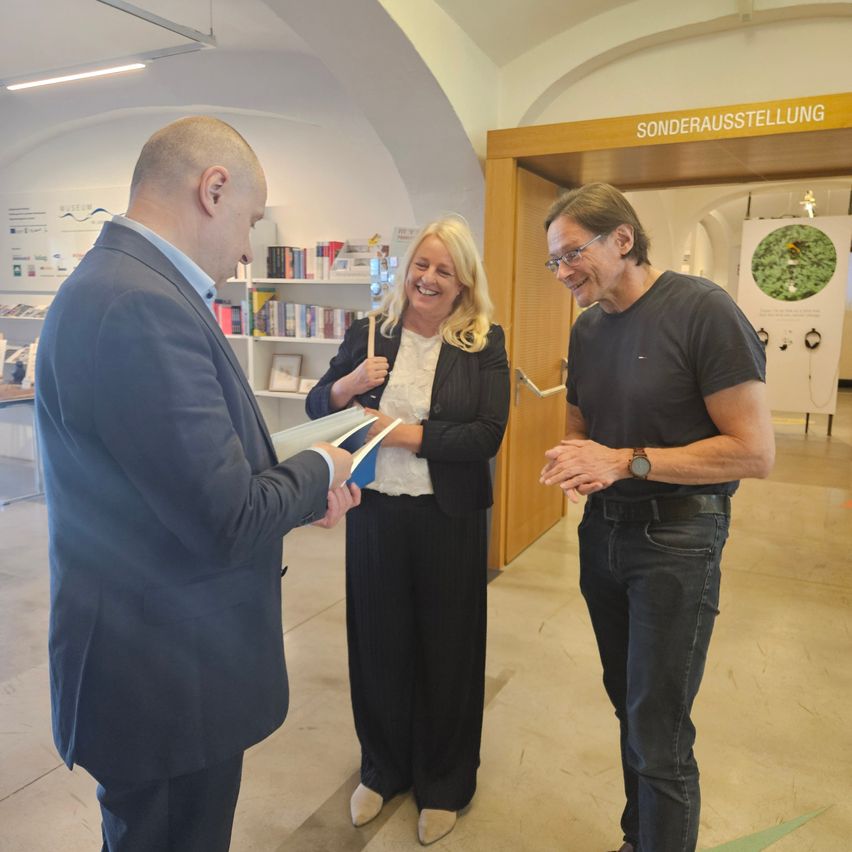 Drei Menschen stehen in einem Raum. Ein Mann hält ein Buch und spricht mit einer Frau. Hinter ihnen befindet sich eine Tür mit einem Schild, das 'Sonderausstellung' steht.
