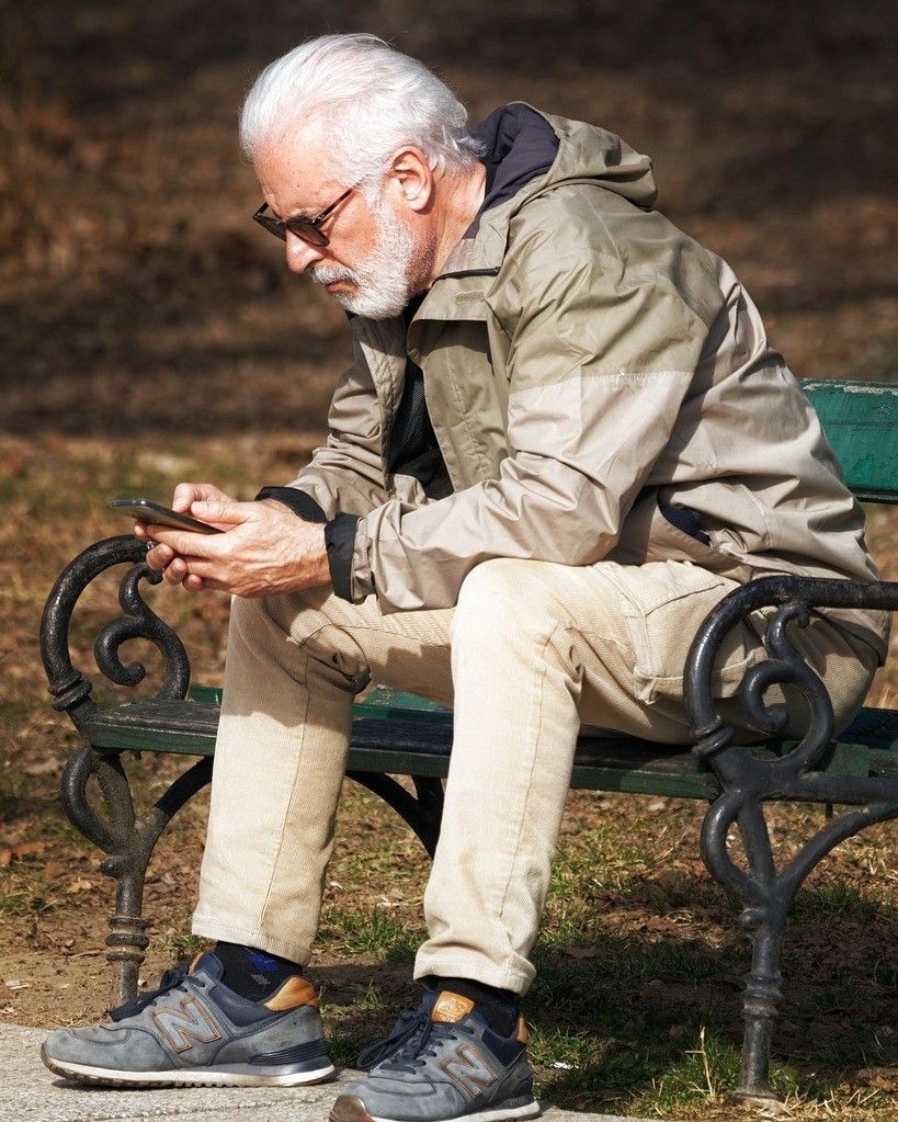 Ein älterer Mann mit Brille und Jacke sitzt auf einer Parkbank und schaut auf sein Smartphone.
