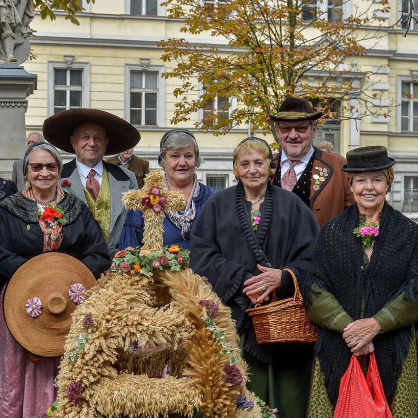 Eine Gruppe älterer Personen in traditioneller Kleidung, die lächelnd und vor einem Gebäude und einer dekorativen Maisstruktur für ein Foto posieren.