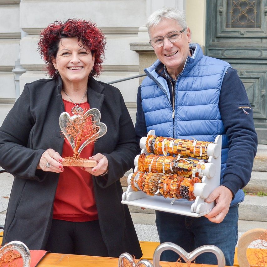 Eine Frau und ein Mann lächeln und posieren für ein Foto. Die Frau hält ein silbernes herzförmiges Objekt und der Mann hält eine Ausstellung von Mais-Armbändern.