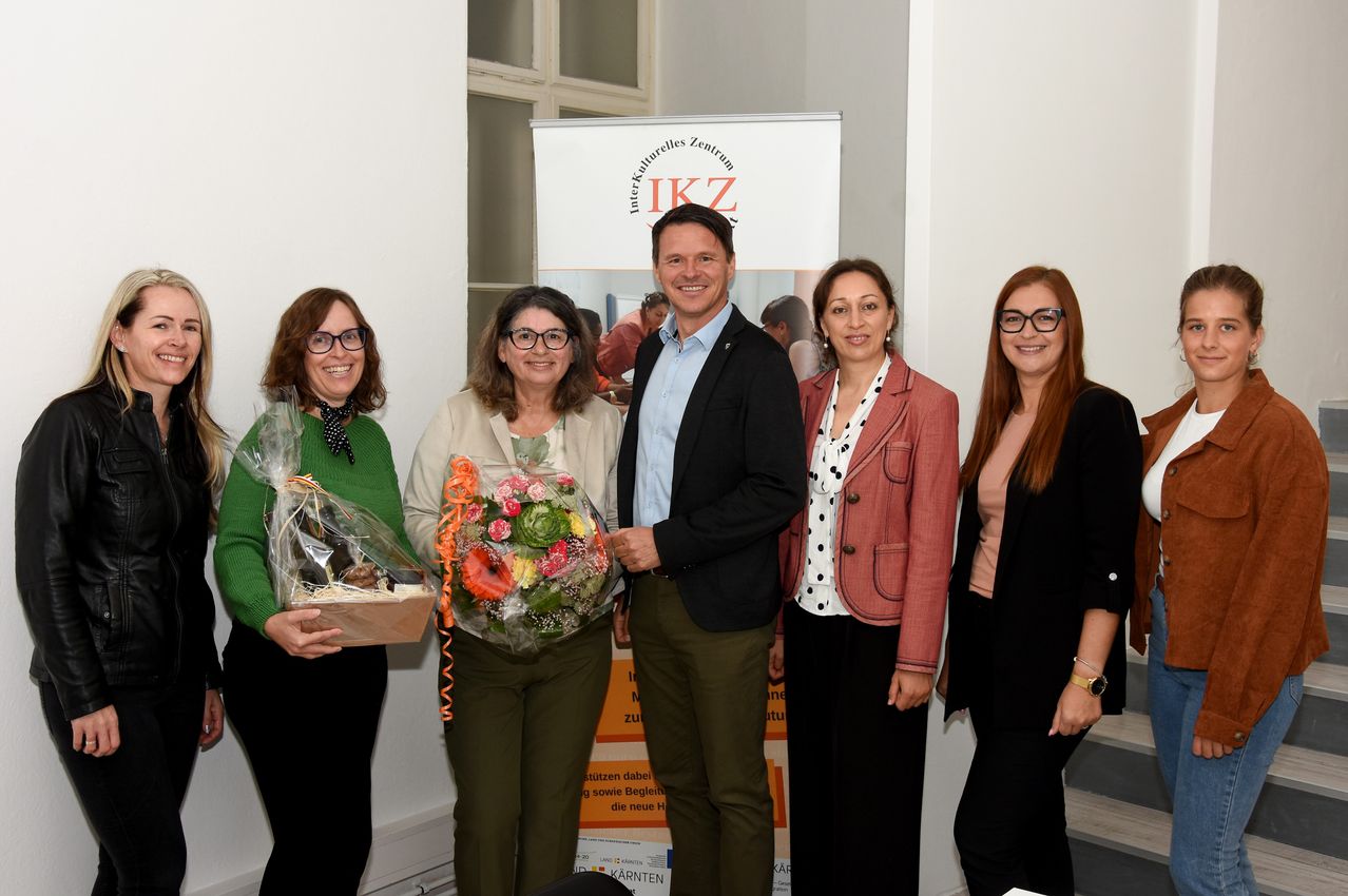 Fünf Personen stehen vor einem Banner mit den Buchstaben IKZ und lächeln für ein Foto. Ein Mann hält einen Blumenstrauß, während andere Geschenke halten. Die Frau auf der linken Seite trägt eine Brille und einen grünen Pullover. Hinter ihnen befindet sich ein Glasfenster.