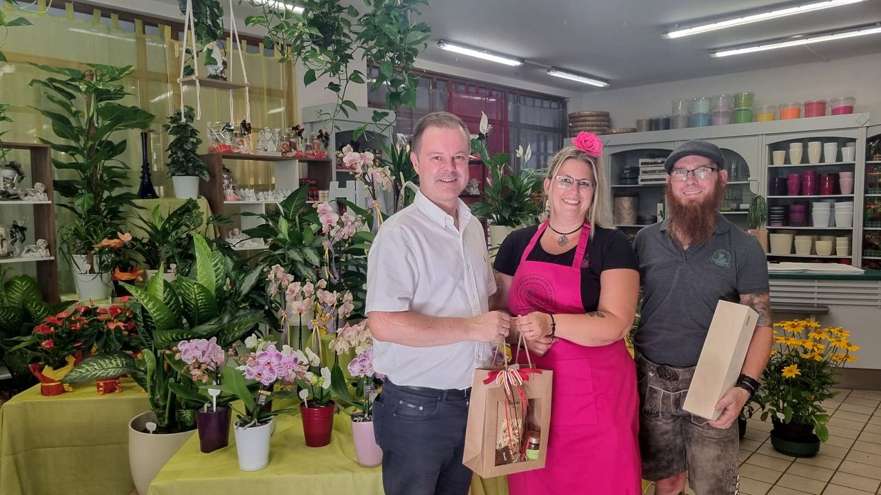 Drei Personen stehen in einem Raum mit vielen Blumen. Ein Mann in einem weißen Hemd übergibt einer Frau in einem rosa Kleid eine Tasche. Ein Mann in einem grauen Hemd steht neben ihnen.
