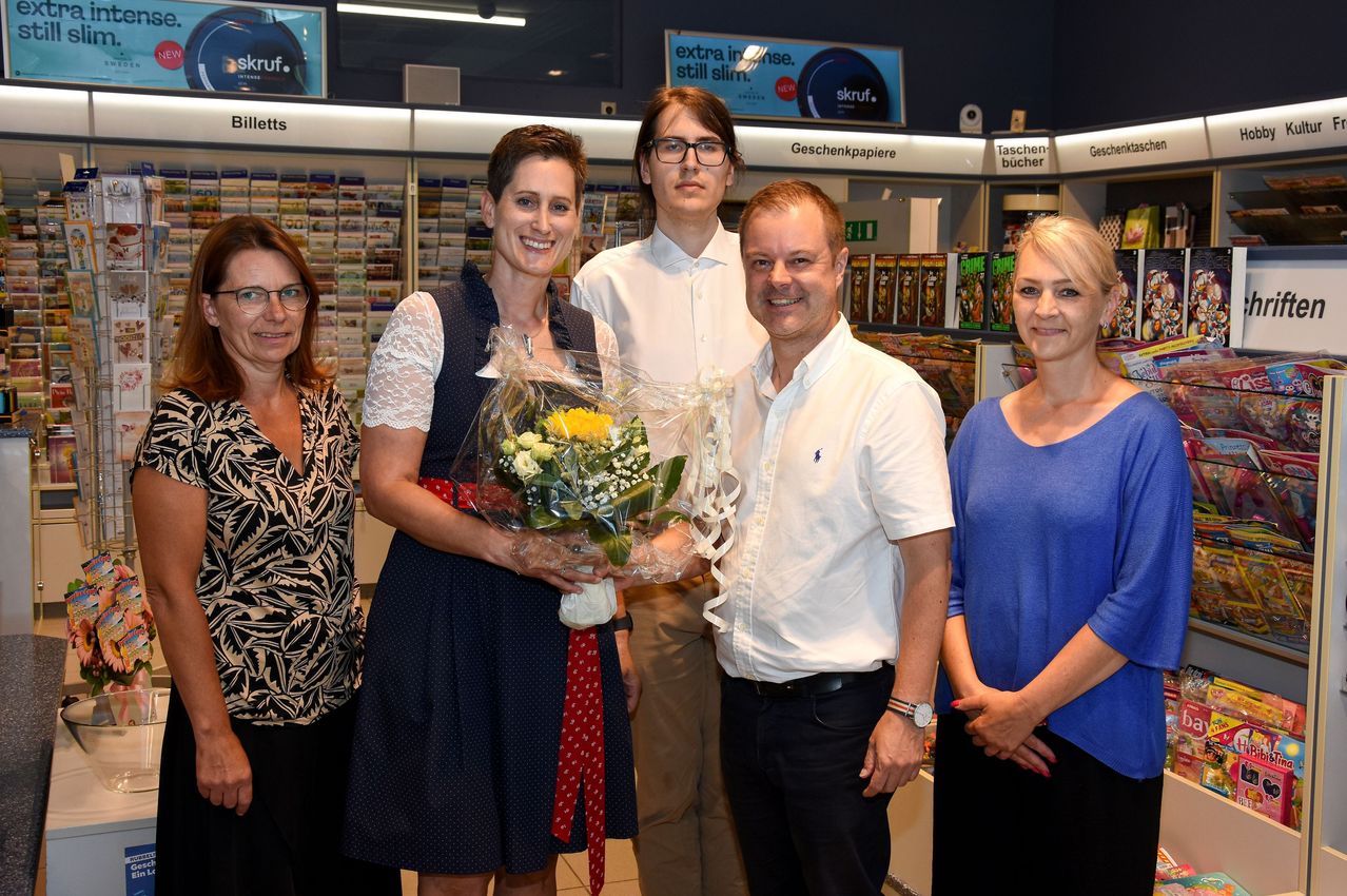 Fünf Personen stehen in einem Geschäft. Eine Frau in einem blauen Kleid hält einen Blumenstrauß. Der Mann neben ihr trägt eine Brille und ein weißes Hemd. Ein weiterer Mann in einem weißen Hemd steht zwischen ihnen. Zwei Frauen an den Seiten lächeln.