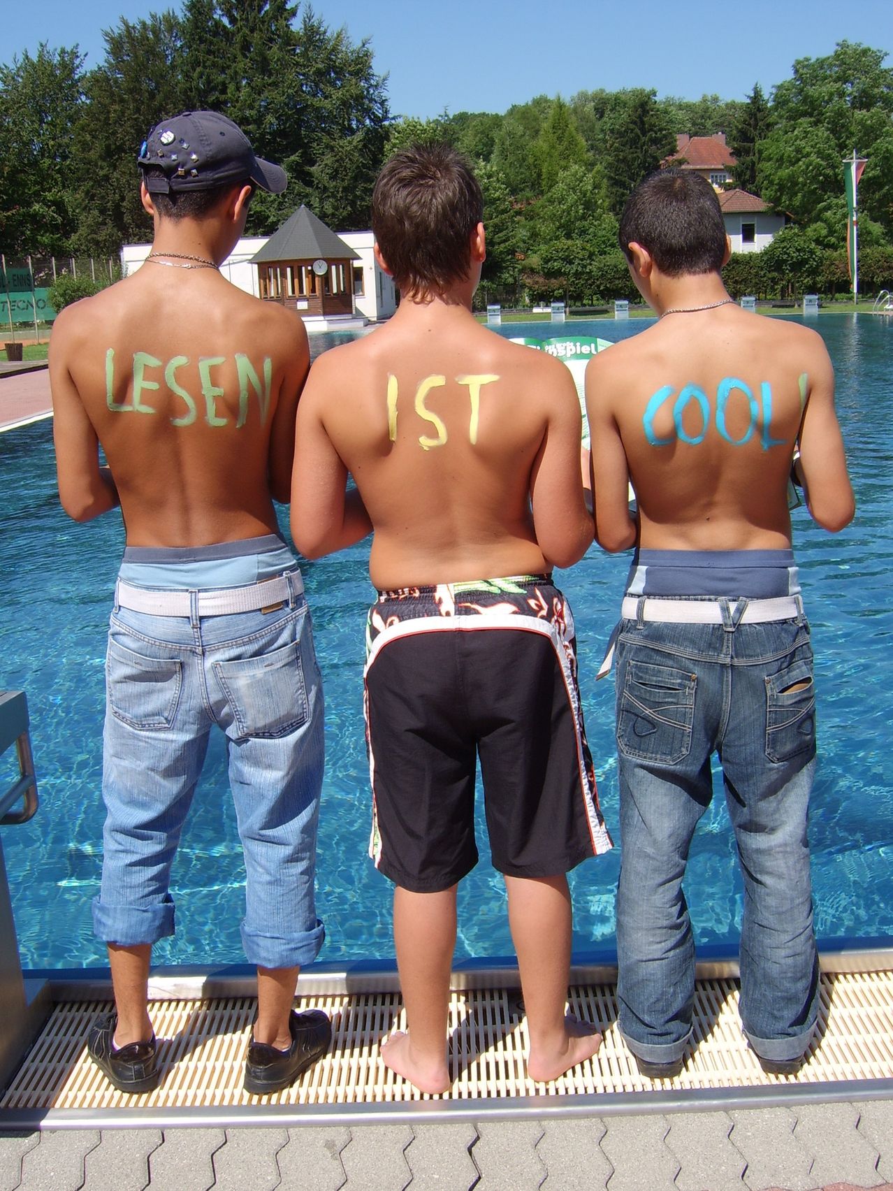 Drei junge Männer stehen mit dem Rücken zur Kamera in der Nähe eines Schwimmbeckens. Sie haben Wörter auf ihren Rücken geschrieben: 'LESEN', 'IST' und 'COOL!'. Sie tragen Jeansshorts und -shirts. Das Becken hat ein Metallgeländer und ist von Bäumen umgeben.