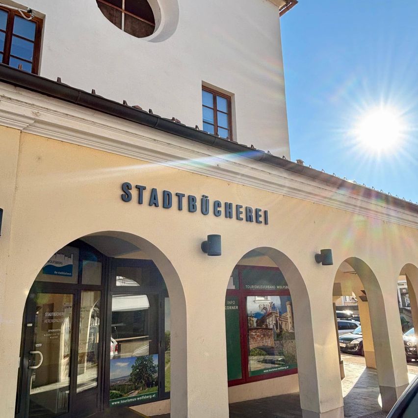 Das Äußere eines weißen Gebäudes mit gewölbten Eingangsbereichen und einem Schild, das 'Stadtbücherei' steht. Das Gebäude hat Glasfenster und einen sonnigen Himmel darüber.