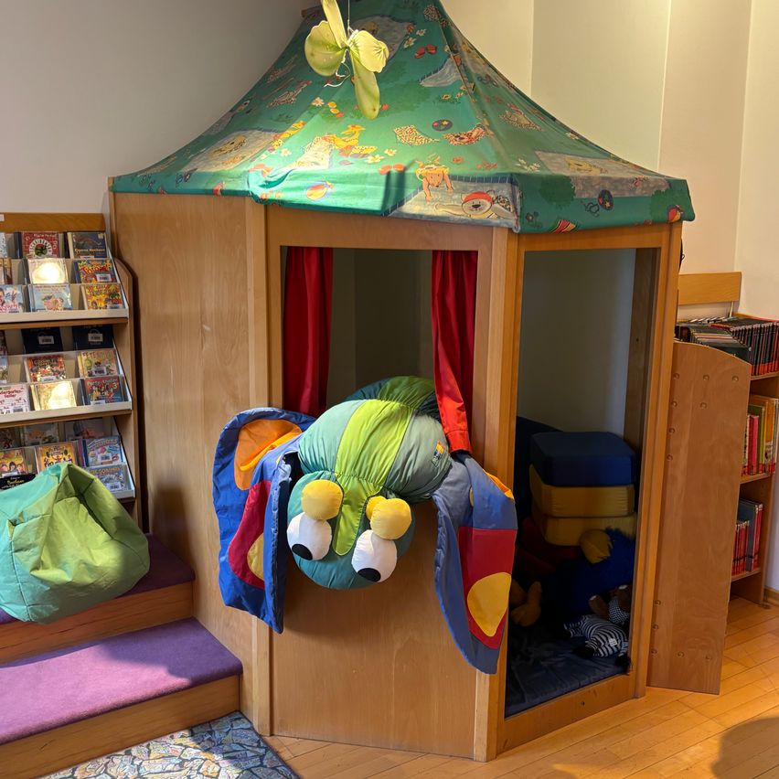 Eine farbenfrohe Leseecke mit einem Zeltdach, einer Plüsch-Raupe und Regalen voller Bücher. Die Ecke ist von Sitzsäcken und einem Teppich umgeben.