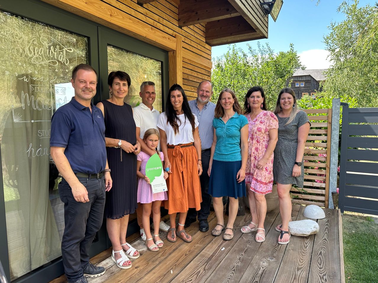 Eine Gruppe von Menschen posiert für ein Foto auf einer Holzveranda mit einem Holzzaun und Pflanzen im Hintergrund.