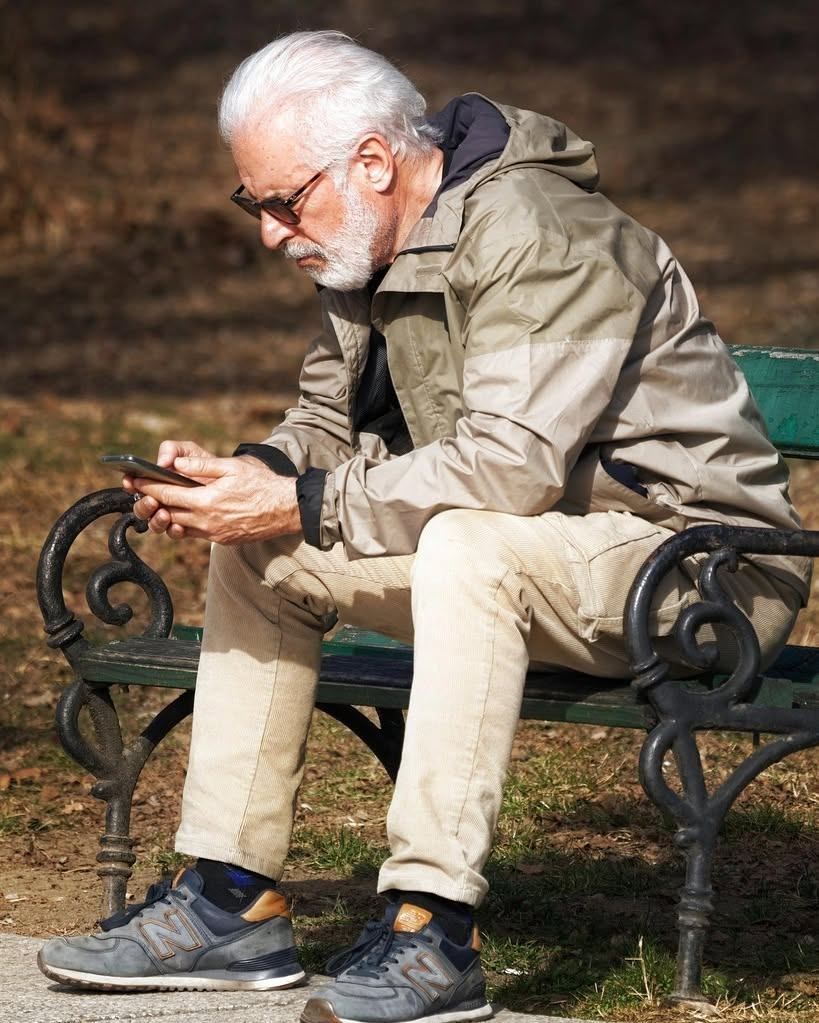 Ein älterer Mann mit Brille und Jacke sitzt auf einer Bank und schaut auf sein Handy.