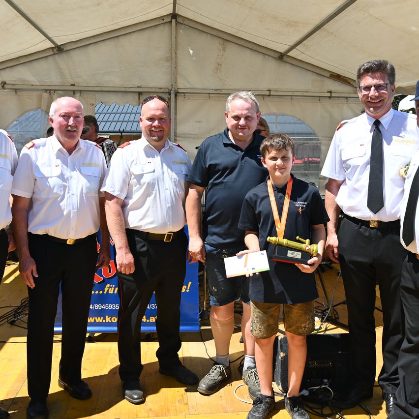 Ein junger Junge mit einer Medaille steht unter einem Zelt zwischen fünf Männern in Uniform, lächelt und hält ein Zertifikat und einen Preis in der Hand.