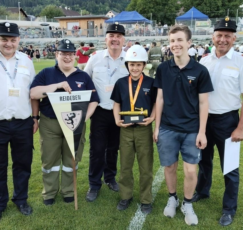 Fünf Erwachsene und ein Kind stehen auf einem Feld und lächeln für ein Foto. Das Kind hält eine Trophäe. Zwei Erwachsene tragen Hüte und Ausweise. Hinter ihnen steht ein Banner mit der Aufschrift 'Reideben'. In der Ferne sind Zelte und eine Menschenmenge zu sehen.