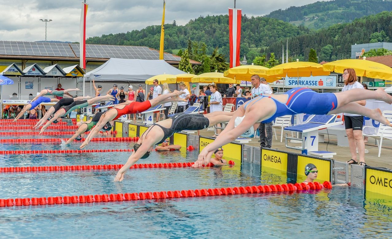 Ein Schwimmwettbewerb mit mehreren Tauchern, die ins Becken springen. Zuschauer und Offizielle sind anwesend. Das Becken hat Bahnmarkierungen und Banner. Im Hintergrund befinden sich Berge und Bäume.