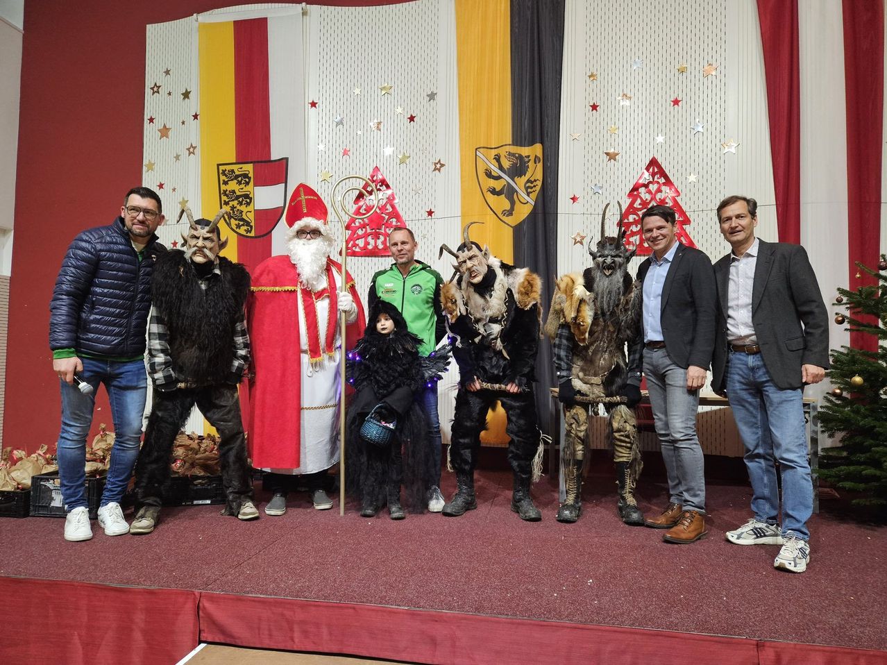 Eine Gruppe von Menschen in verschiedenen Kostümen steht auf einer Bühne. Ein Mann in einem Weihnachtsmannkostüm steht in der Mitte. Zwei Menschen in Hörnerkostümen stehen zu seinen Seiten. Dahinter stehen drei Menschen.