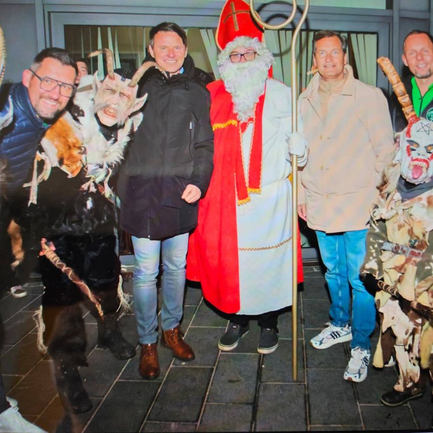 Fünf Männer stehen in einer Reihe. Einer ist als Weihnachtsmann verkleidet, während die anderen verschiedene Kostüme tragen. Sie alle lächeln und scheinen für ein Foto zu posieren.