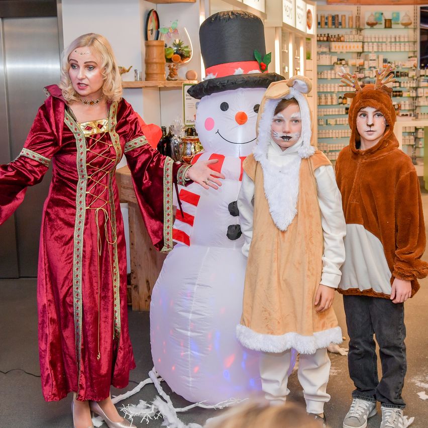 Eine Frau in einem roten Kostüm steht neben einem großen aufblasbaren Schneemann. Zwei Kinder, verkleidet als Rentier und Eisbär, stehen neben dem Schneemann.