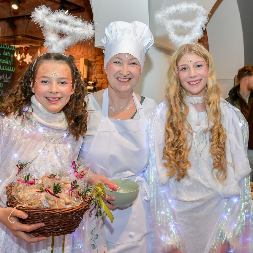 Zwei Mädchen und eine Frau in weißen Kostümen, eine hält einen Korb mit Essen und die andere einen Schüssel, stehen zusammen lächelnd. Die Frau trägt einen Kochmütze und eine Schürze. Hinter ihnen ist ein verschwommener Hintergrund mit einer Person.