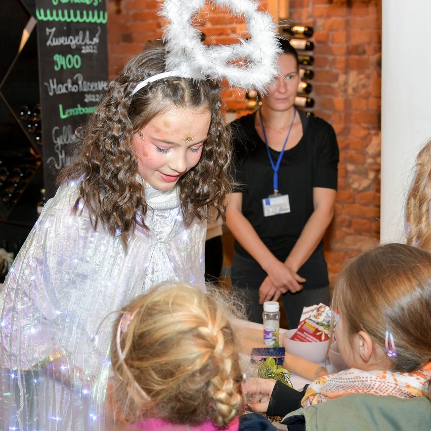 Ein junges Mädchen mit Engelsflügeln interagiert mit Kindern in einem Raum. Hinter ihr steht eine Frau in einem schwarzen Shirt an einer Ziegelwand. Es ist ein Schild und Weinflaschen sichtbar.