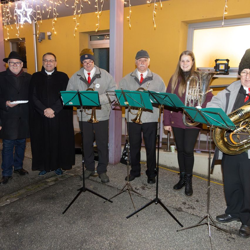 Sechs Personen, darunter vier Männer und zwei Frauen, stehen hinter Notenständern mit Blechblasinstrumenten. Sie sind in Winterkleidung gekleidet und scheinen zu spielen. Die Szene findet draußen bei Nacht statt.