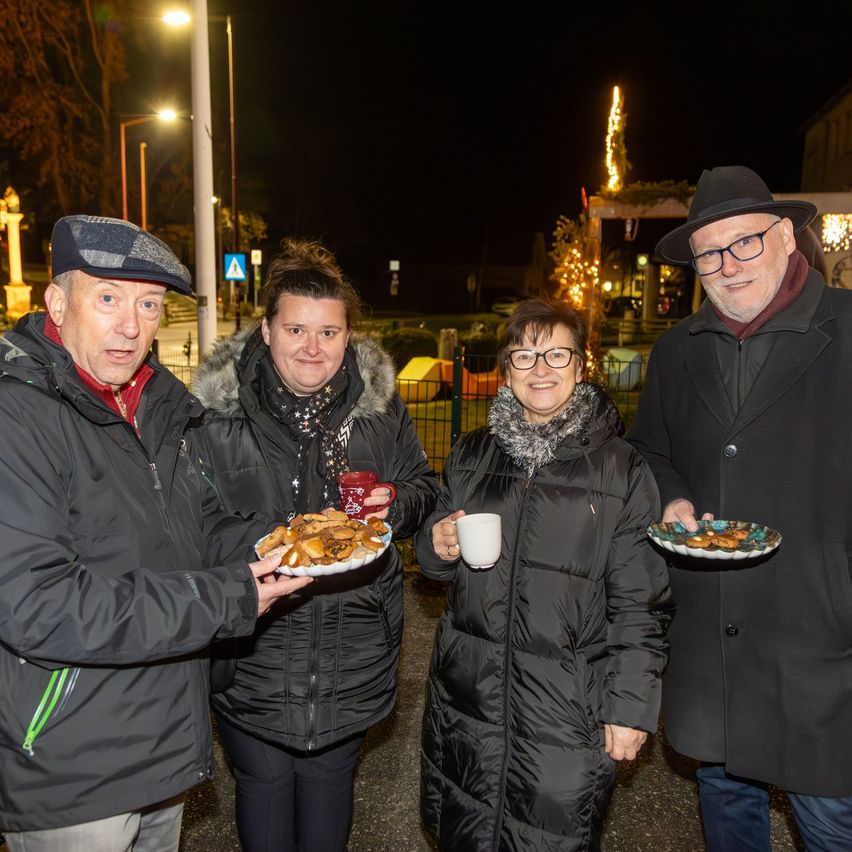 Vier Menschen posieren nachts im Freien für ein Foto, halten Teller mit Essen in den Händen. Die Frau in der Mitte hält eine Tasse. Sie tragen alle Wintermäntel. Im Hintergrund sind Straßenlaternen und eine dekorative Lichterkette zu sehen.