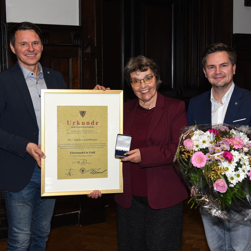 Drei Personen stehen lächelnd zusammen. Die Frau in der Mitte hält ein gerahmtes Zertifikat und eine Medaille. Der Mann auf der linken Seite hält den Rahmen. Der Mann auf der rechten Seite hält einen Blumenstrauß. Der Hintergrund wirkt feierlich.