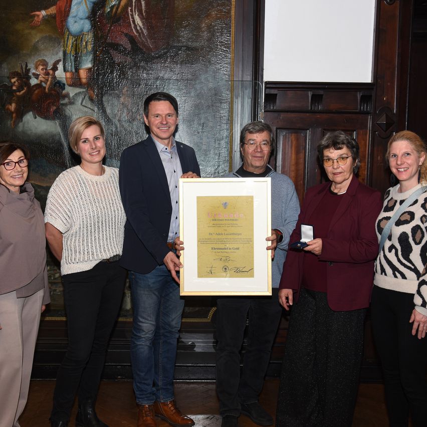 Fünf Menschen stehen lächelnd drinnen, ein großes gerahmtes Zertifikat, das von zwei Männern in der Mitte gehalten wird. Dahinter ist ein großes Wandbild.