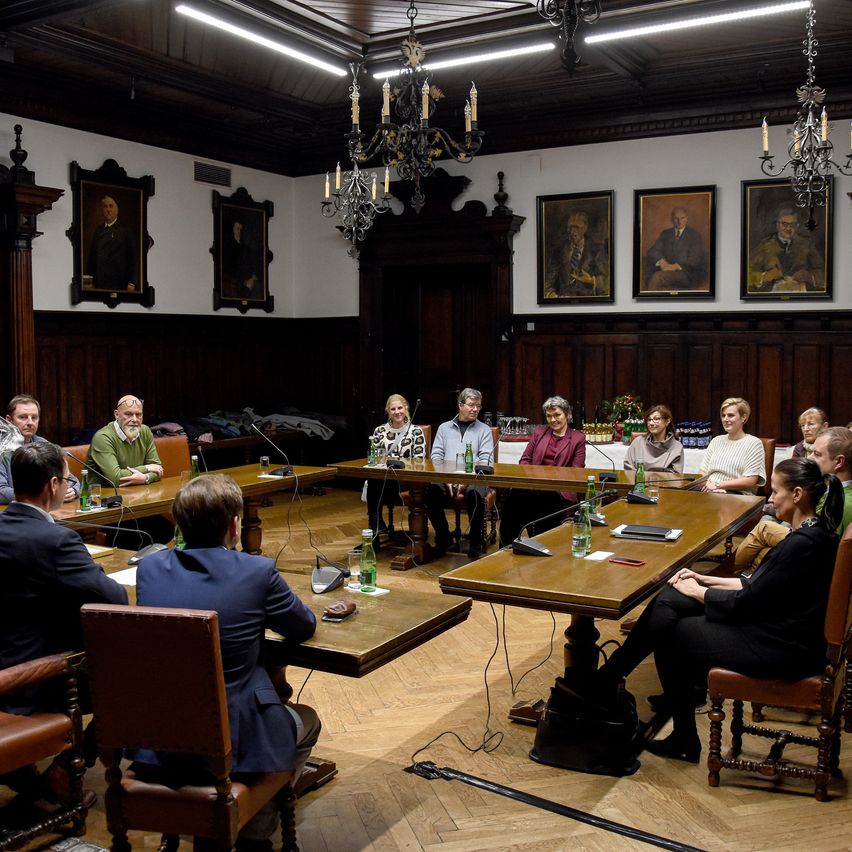 Ein Konferenzraum mit Holzvertäfelung und Kronleuchtern, eine Gruppe von Menschen sitzt um einen langen Tisch mit Mikrofonen, Wasserflaschen und Papieren, mit mehreren gerahmten Porträts an der Wand.