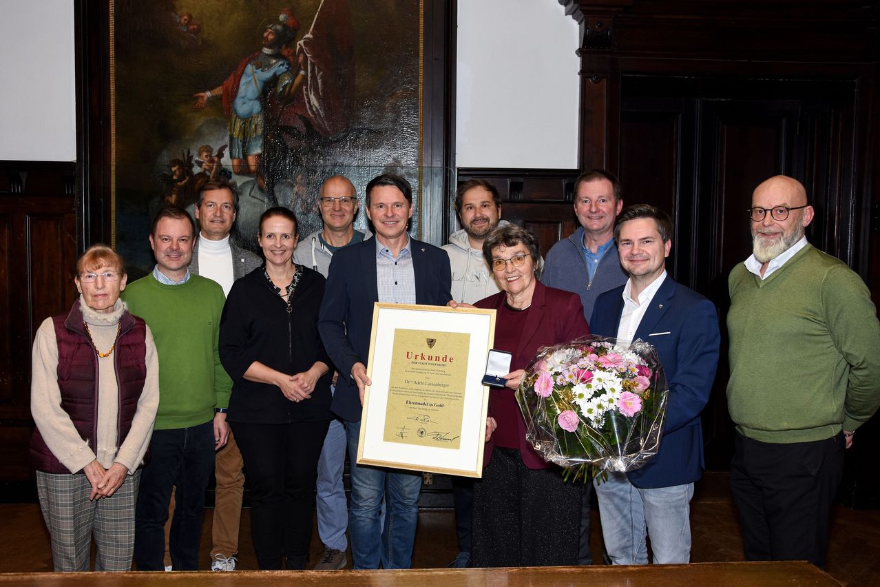 Eine Gruppe von Menschen steht lächelnd in einer formellen Umgebung. Eine Frau hält ein gerahmtes Zertifikat und einen Blumenstrauß. Hinter ihnen schmückt ein Gemälde die Wand.