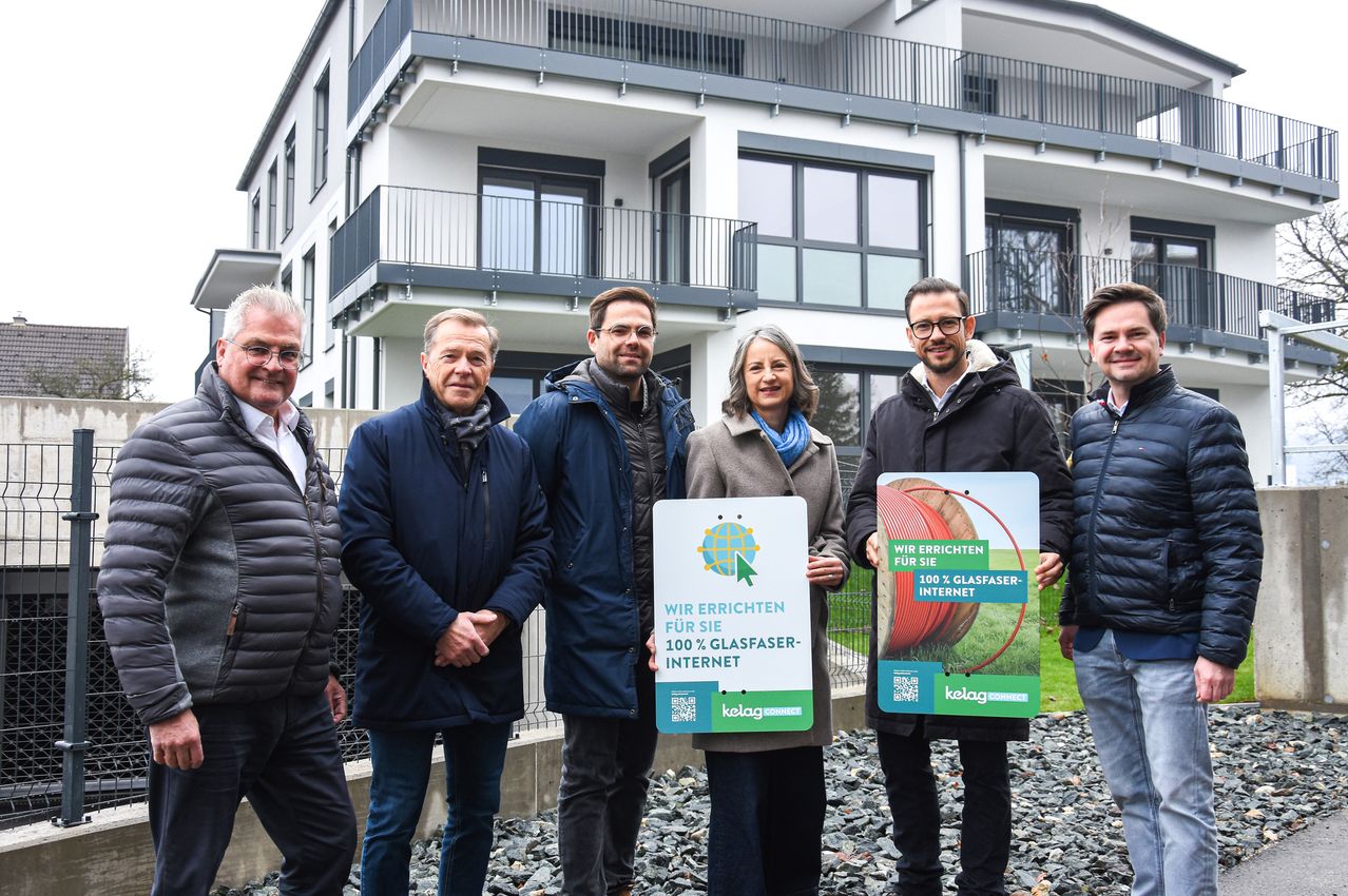 Fünf Personen, zwei Männer auf jeder Seite und eine Frau in der Mitte, stehen vor einem modernen Gebäude. Sie halten Schilder mit der Aufschrift 'Wir errichten 100% Glasfaser-Internet'.