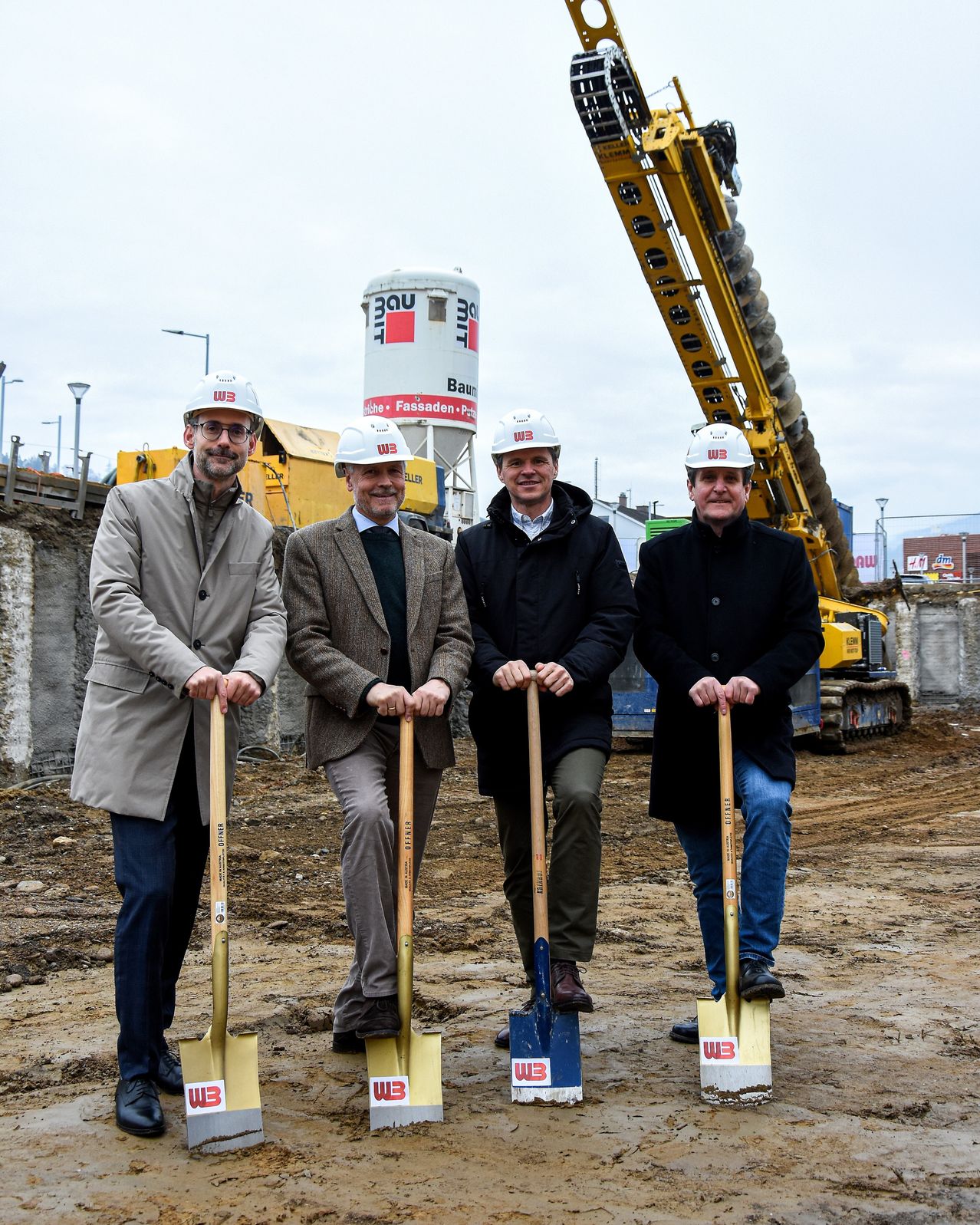 Vier Männer in weißen Schutzhelmen halten Schaufeln an einer Baustelle, mit einem großen gelben Bagger im Hintergrund.