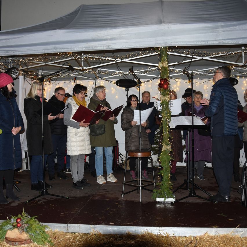A choir performs under a white tent with holiday decorations, microphones, and music stands, while people in winter clothing listen.