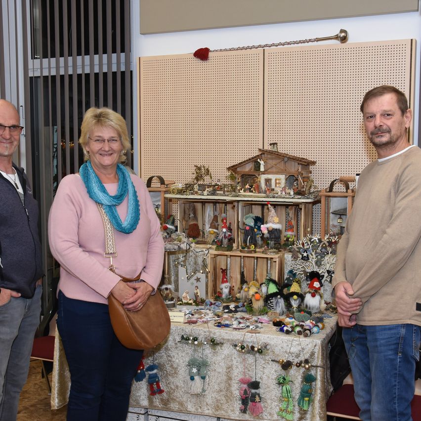 Three people stand in front of a display table with Christmas decorations, including a model house, figurines, and ornaments. The woman wears a pink sweater and a blue scarf. The man on the right has a beard and wears a beige shirt.