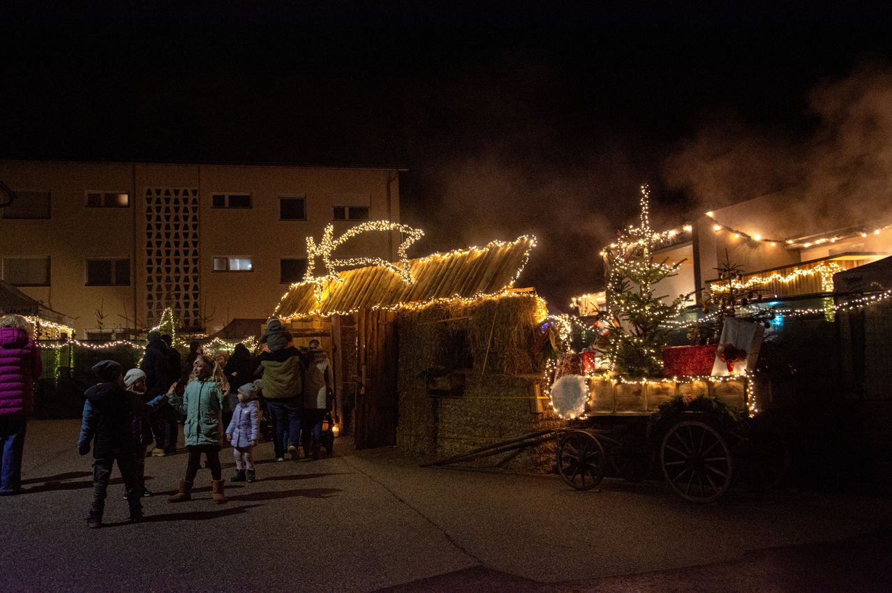 A decorated nativity scene with lights, straw roof, and a small Christmas tree stands in front of a building. Children and adults gather around, enjoying the festive atmosphere.