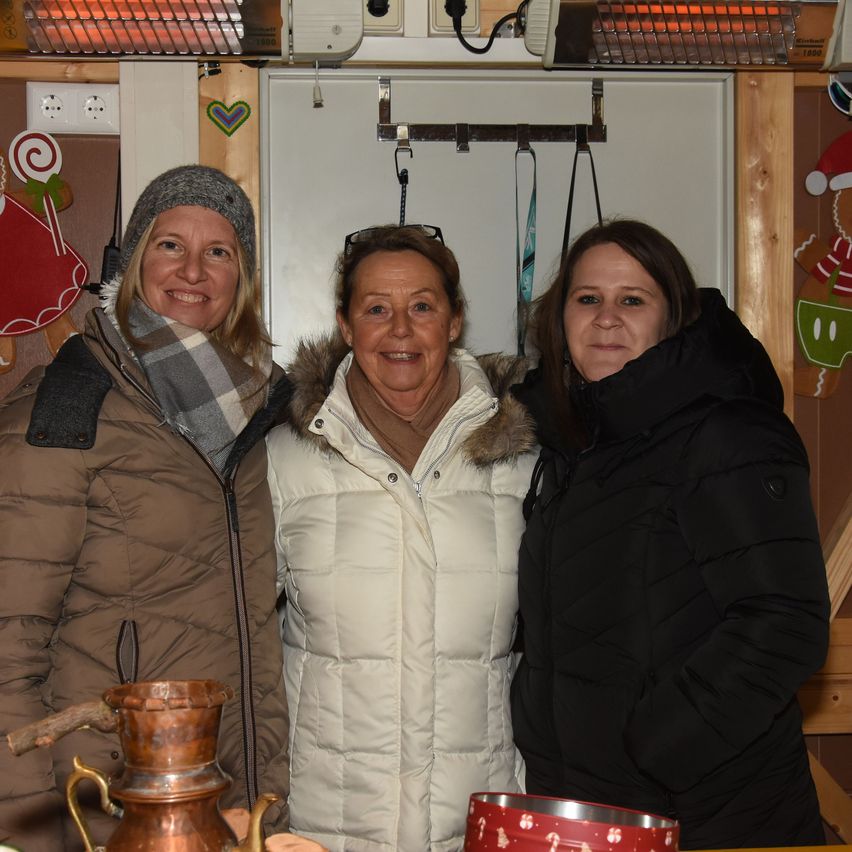 Three women are standing together in a room, smiling for a photo. One wears a beanie and scarf, another has glasses on her head, and the third is in a black jacket. Behind them, a Christmas decoration and a heater are visible.