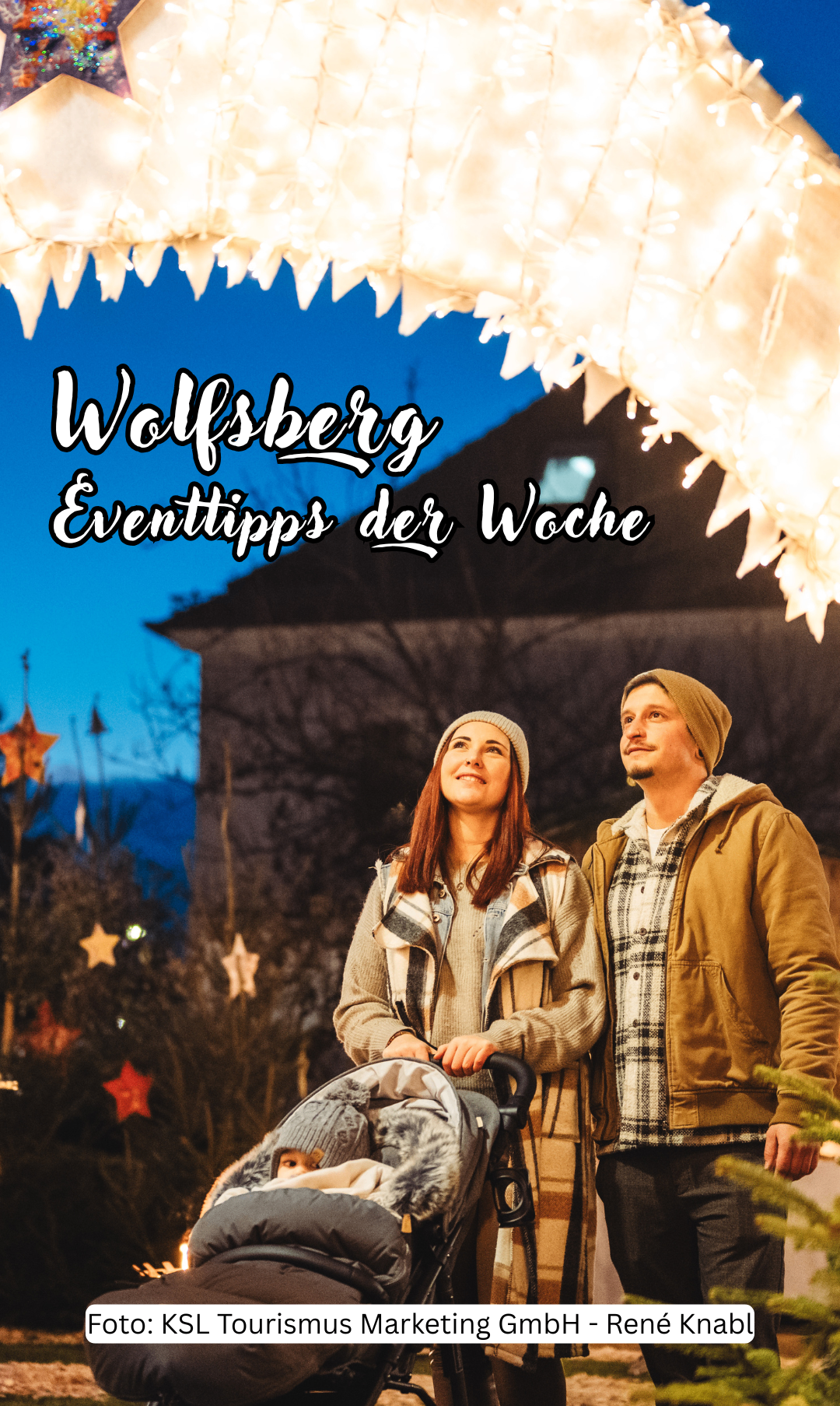 A couple stands outside with a stroller, smiling at the camera, with illuminated decorations and a building in the background.