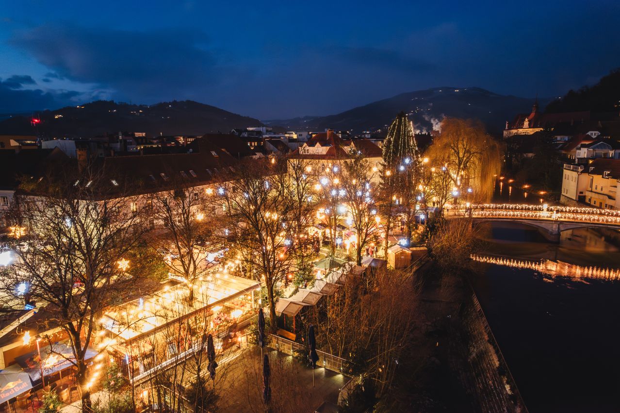 Eine Stadt bei Nacht mit Gebäuden, Bäumen und Lichtern. Es gibt eine Brücke über einem Fluss mit Lichtreflexionen. Berge sind in der Ferne.