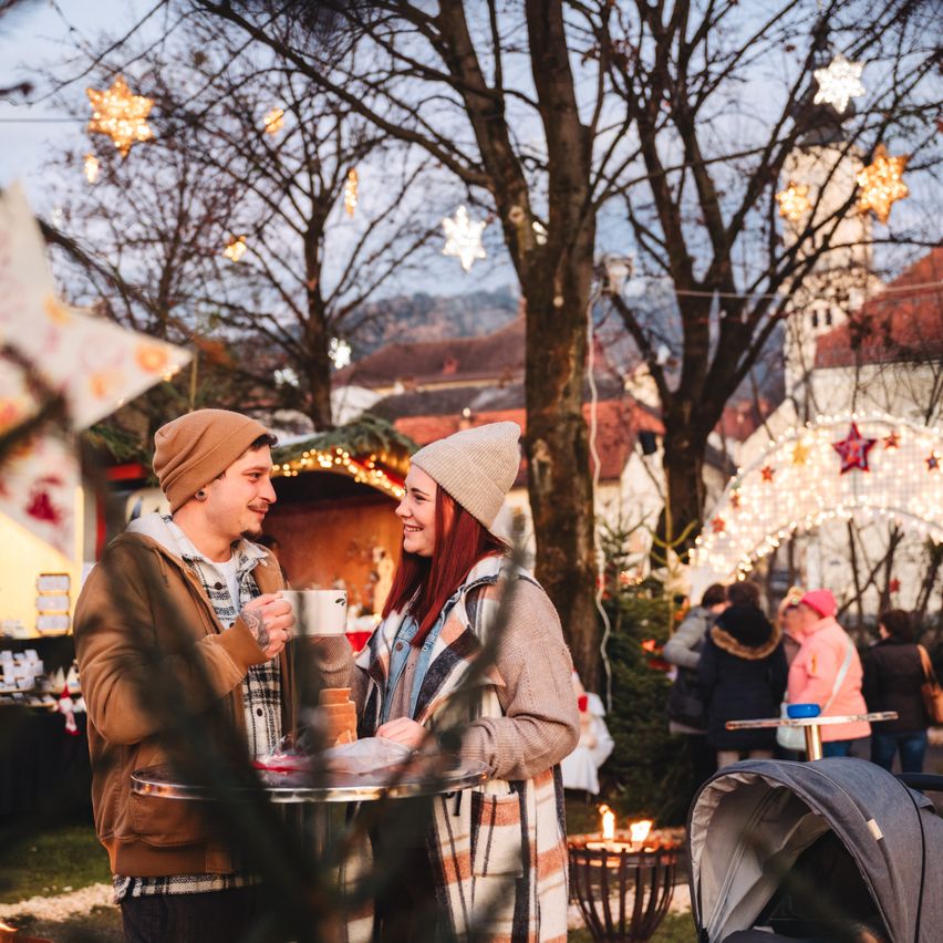 Ein Paar steht bei einem Tisch, genießt einen Moment, hält Tassen in einem festlichen Weihnachtsmarkt mit Bäumen und Dekorationen. Im Hintergrund sind andere Leute und ein Kinderwagen zu sehen.