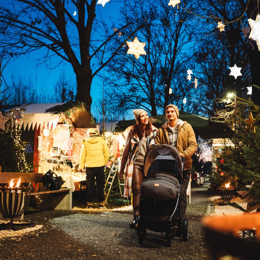 Ein Paar läuft nachts durch einen festlichen Markt und geht an einem Kinderwagen vorbei, vorbei an Geschäften, Bäumen und Lichtern, die den Bereich schmücken.