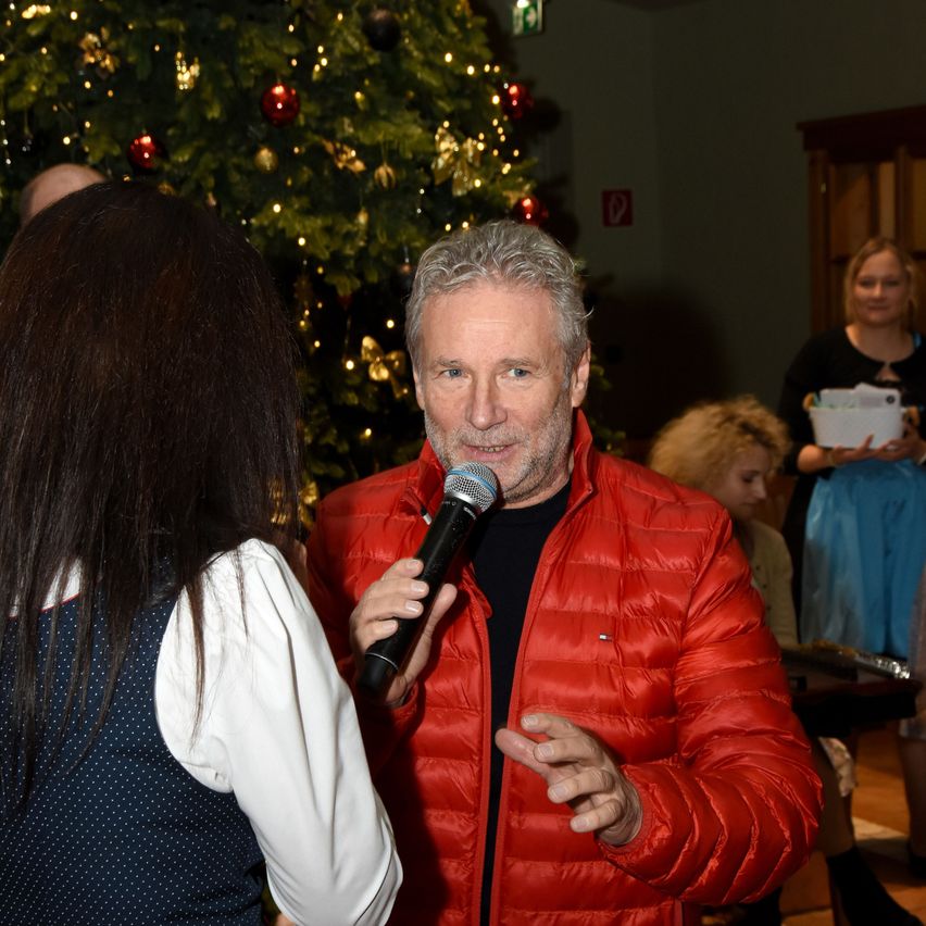 Ein Mann mit Mikrofon spricht hinein, während ein aufmerksamer Zuhörer zuhört, vor dem Hintergrund eines festlich geschmückten Weihnachtsbaums.