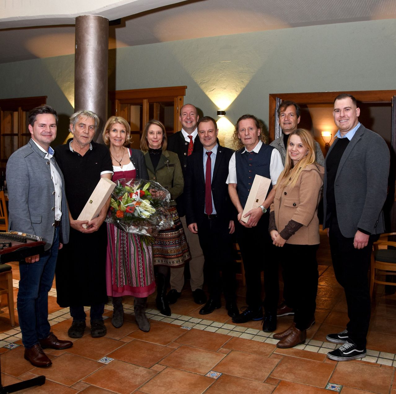 Eine Gruppe von Menschen, einige halten Auszeichnungen und Blumensträuße, stehen in einem Raum mit gefliesten Böden und Holztüren. Sie scheinen ein Ereignis zu feiern.