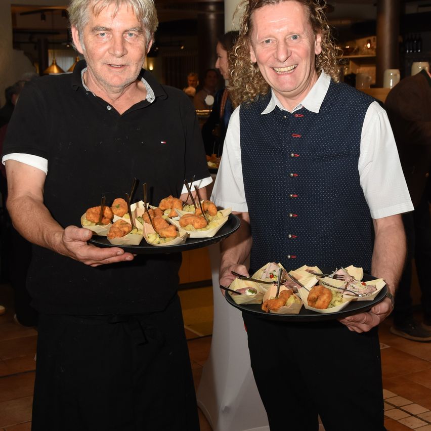 Zwei Männer stehen in einem Raum und halten Tabletts mit Essen. Der Mann auf der linken Seite trägt ein schwarzes Shirt und der Mann auf der rechten Seite trägt eine Weste.