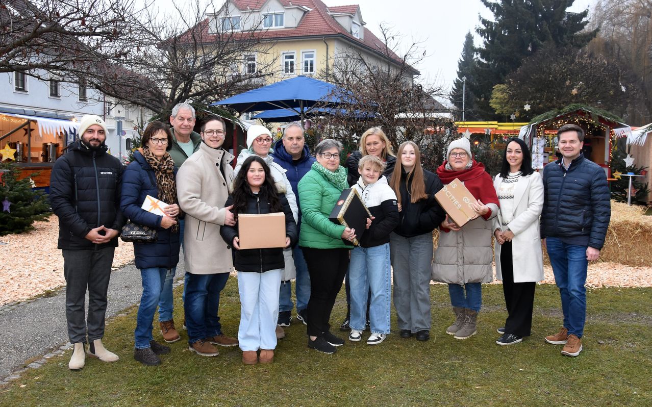 Eine Gruppe von Erwachsenen und Kindern steht auf einem Rasen und hält Kisten, wahrscheinlich für ein Foto. Dahinter befinden sich Gebäude und Bäume.