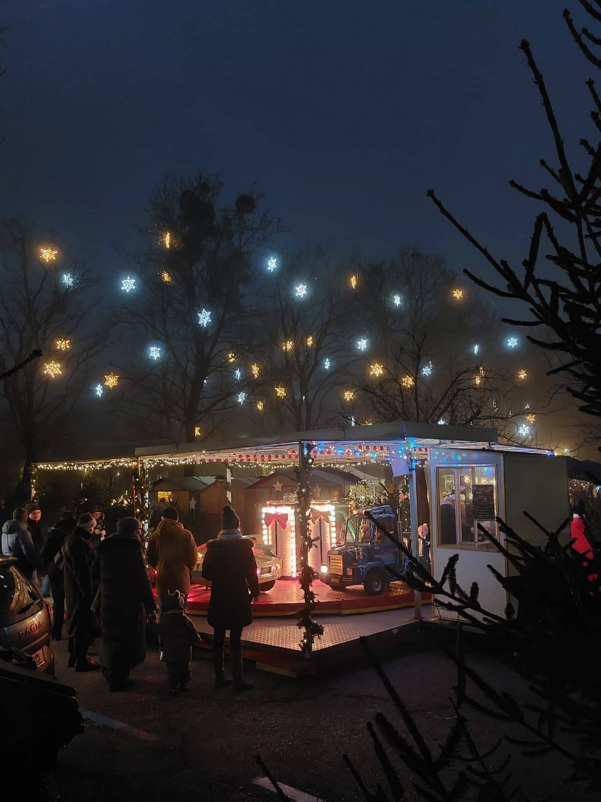 Eine Nachtszene eines Weihnachtsmarktes mit einem Karussell, Menschen in Winterkleidung und sternförmigen Lichtern, die von Bäumen hängen.