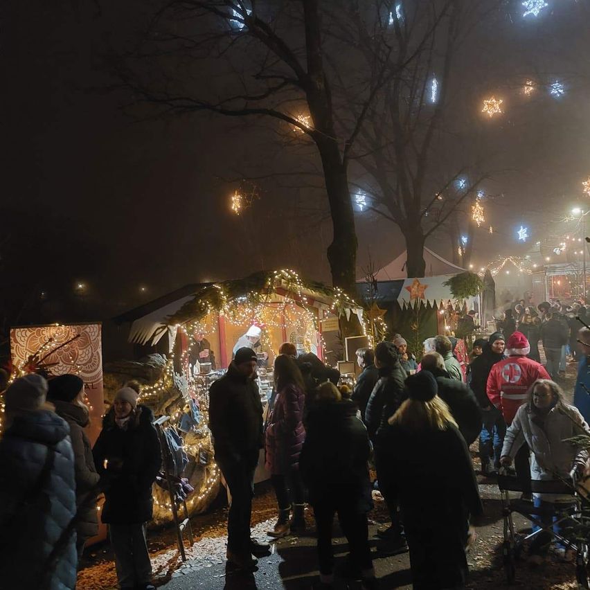 Ein Weihnachtsmarkt bei Nacht ist voller Menschen. Stände sind mit bunten Lichtern und Dekorationen geschmückt. Leute stöbern durch die Stände, während andere plaudern. Straßenlaternen und dekorative Lichter tragen zur festlichen Atmosphäre bei.