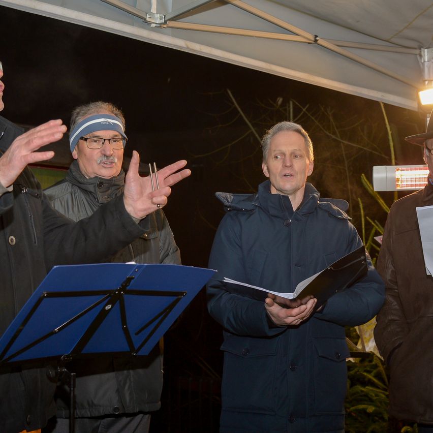 Vier Männer stehen unter einem Zelt, einer von ihnen gestikuliert mit den Händen. Der Mann in der Mitte hält ein Musikbuch. Der Mann auf der rechten Seite trägt einen Hut und hält ein Stück Papier.