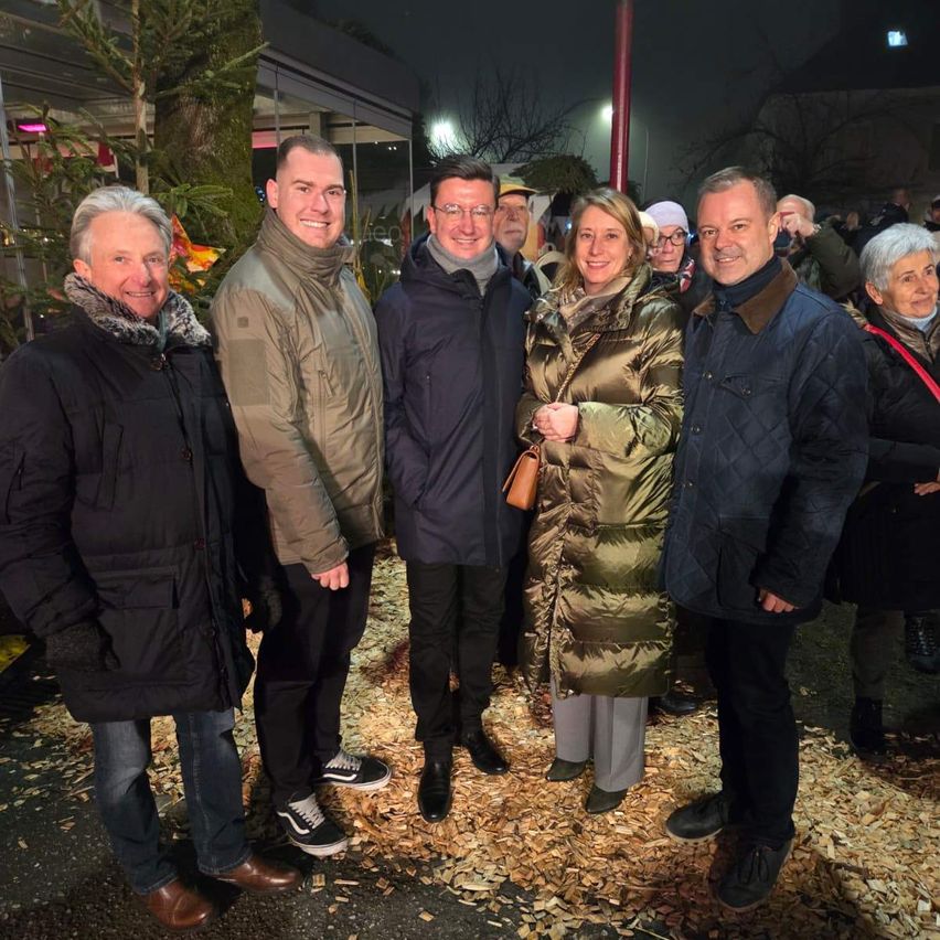 Eine Gruppe von Menschen steht nachts vor einem Gebäude. Sie sind alle warm in Wintermänteln gekleidet. Es gibt einen Weihnachtsbaum und eine festliche Atmosphäre.