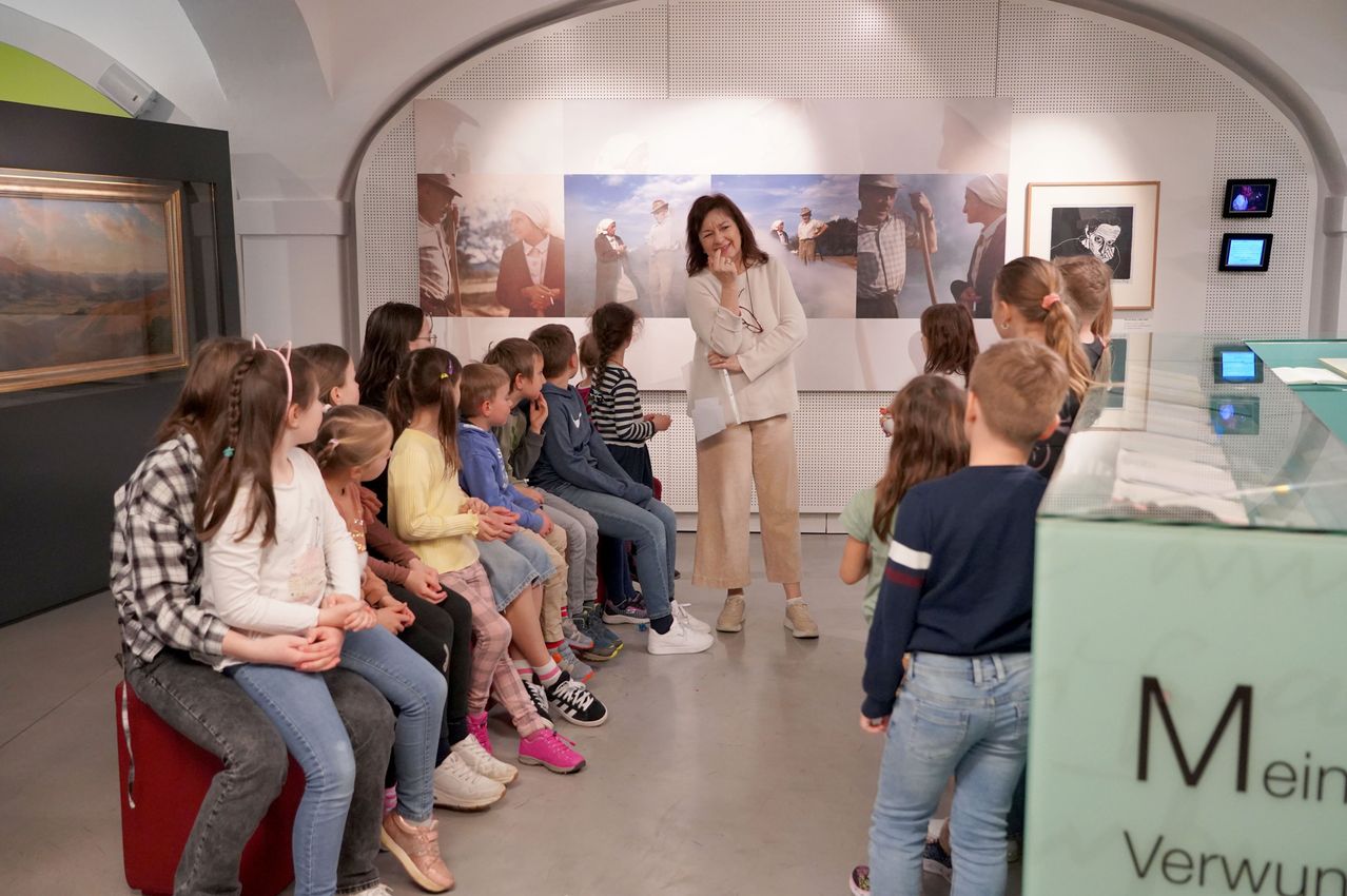 Eine Frau spricht zu einer Klasse von Kindern, die auf einer Bank vor einer Wand mit großen Fotos sitzen.