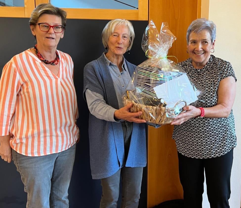 Drei ältere Frauen stehen lächelnd zusammen, wobei eine eine verpackte Geschenktüte hält.