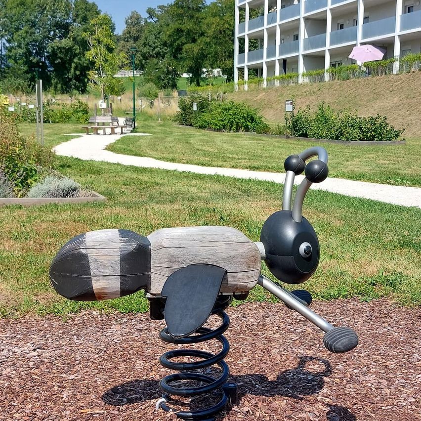 Ein hölzerner Bienenförmiger Federreiter steht auf einem Spielplatz mit Mulchboden, einem Grasfeld und einem Gebäude mit Balkonen in der Ferne.