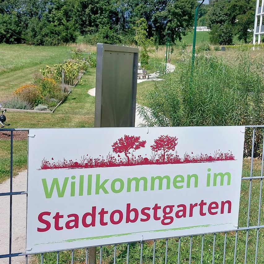 Ein Schild mit den Worten 'Willkommen im Stadtobstgarten' ist an einem Metallzaun in einem Garten befestigt. Der Garten hat eine Vielzahl von Pflanzen und Bäumen. In der Ferne befindet sich ein Gebäude mit weißen Wänden.