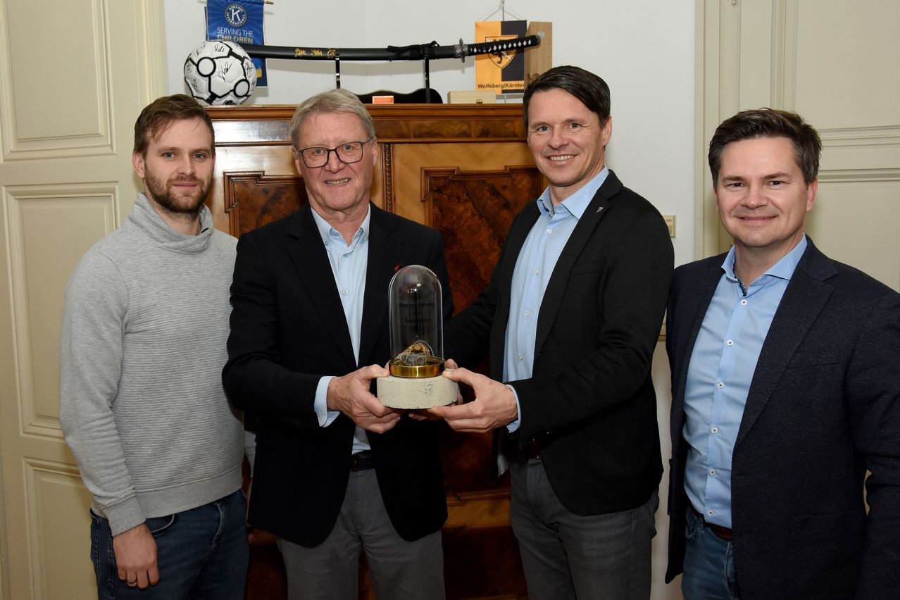 Vier Männer stehen zusammen, zwei halten eine Trophäe, mit einem Holzschrank und Gegenständen im Hintergrund.