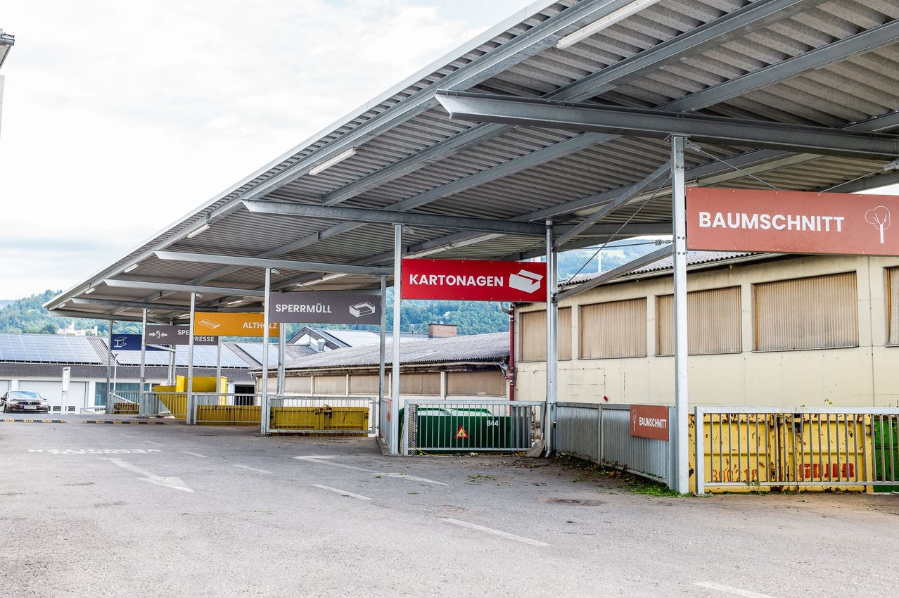 Ein großer überdachter Bereich mit mehreren Schildern, darunter 'KARTONAGEN' und 'BAUM'. Gelbe Müllcontainer und Zäune sind vorhanden. Der Boden ist gepflastert.